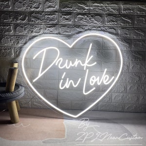 May include: A white neon sign in the shape of a heart with the words "Drunk in Love" in cursive script. The sign is mounted on a white brick wall. The sign is made by ZP ZL Neon Custom.