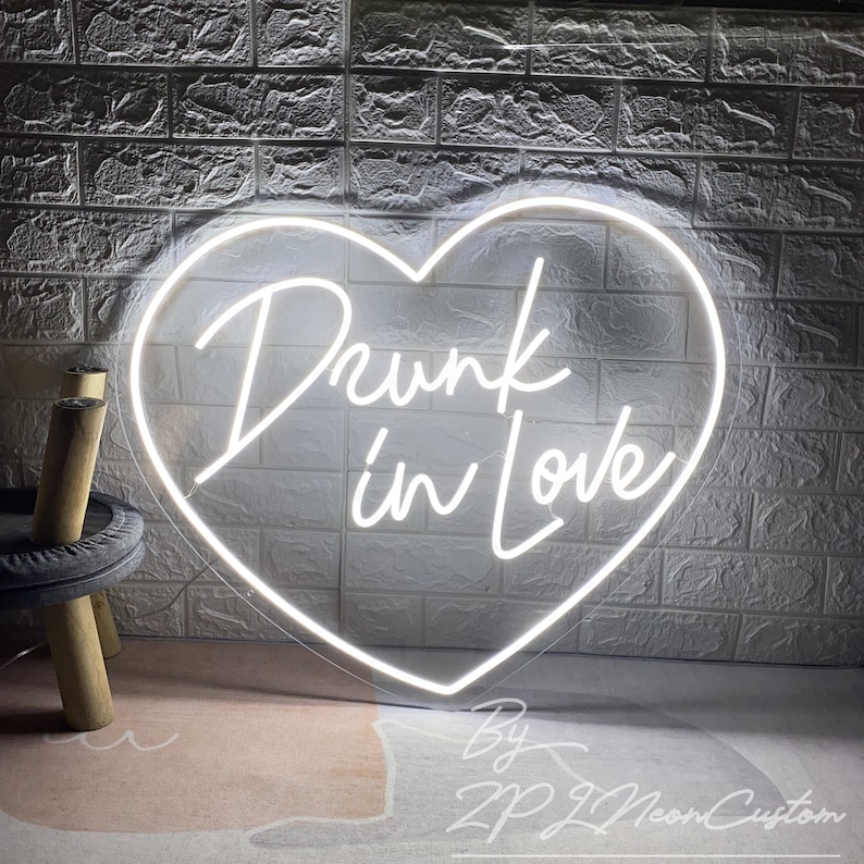 May include: A white neon sign in the shape of a heart with the words "Drunk in Love" in cursive script. The sign is mounted on a white brick wall. The sign is made by ZP ZL Neon Custom.