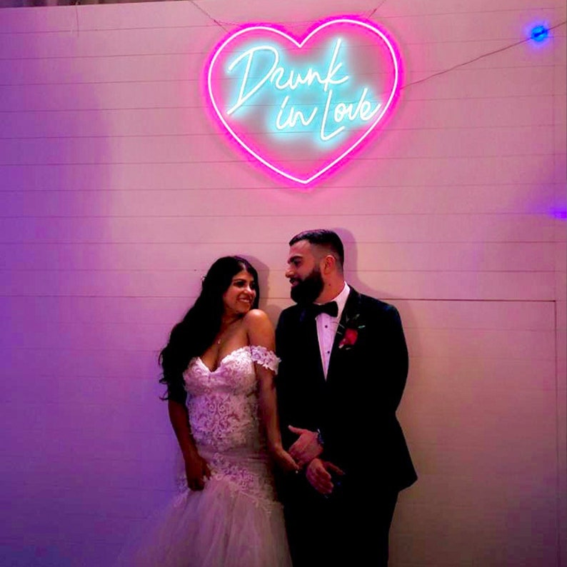 May include: A couple stands in front of a wall with a neon sign. The sign is in the shape of a heart and says "Drunk in Love" in a cursive font. The woman wears a white off-the-shoulder wedding dress. The man wears a black tuxedo.