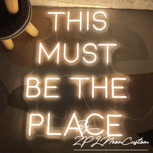 Op de afbeelding: Een neonbord met de tekst "THIS MUST BE THE PLACE" in witte letters. Het bord heeft een rechthoekige vorm en straalt een warm licht uit. Het bord bevat ook de tekst "ZPZL Neon Custom".