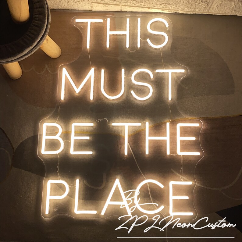 Op de afbeelding: Een neonbord met de tekst "THIS MUST BE THE PLACE" in witte letters. Het bord heeft een rechthoekige vorm en straalt een warm licht uit. Het bord bevat ook de tekst "ZPZL Neon Custom".