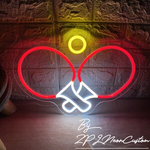 May include: A neon sign depicting a ping pong paddle and ball. The paddles are outlined in red, with white accents, and the ball is yellow. The sign is against a textured wall. Text at the bottom reads "By ZP Neon Custom."
