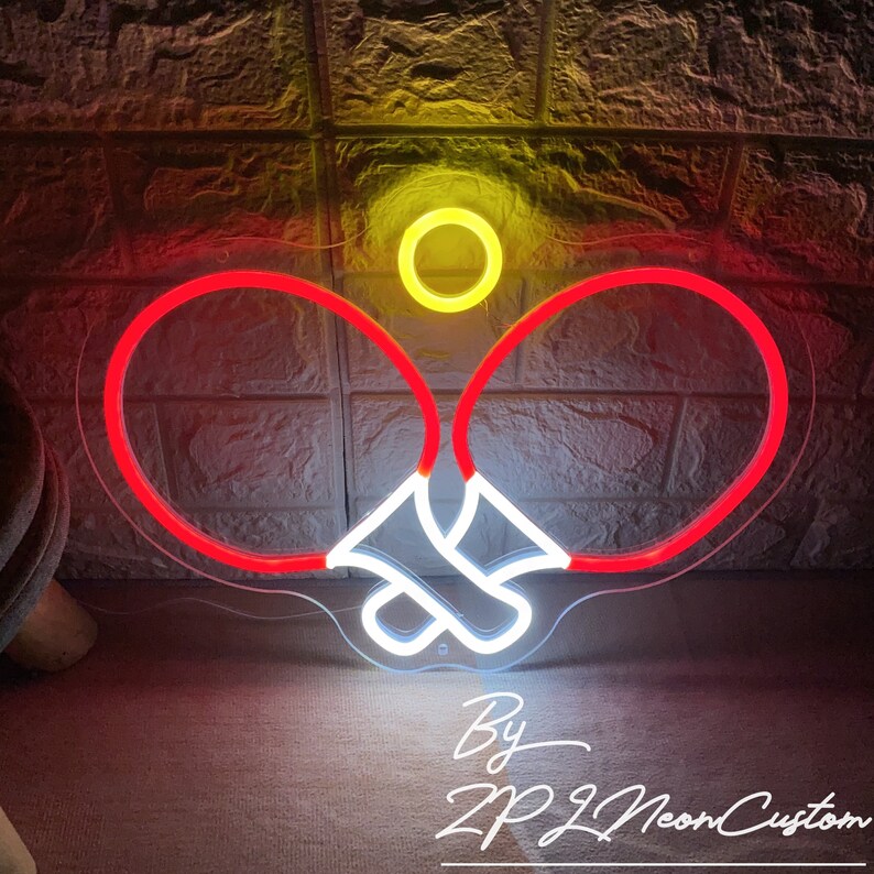 May include: A neon sign depicting a ping pong paddle and ball. The paddles are outlined in red, with white accents, and the ball is yellow. The sign is against a textured wall. Text at the bottom reads "By ZP Neon Custom."