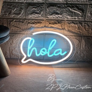 Op de afbeelding: Een neonbord in de vorm van een tekstballon met het woord "hola" in cursief schrift. Het bord heeft een witte omtrek en een blauw interieur en is gemonteerd op een transparante acryl achtergrond. Het bord is verlicht tegen een bakstenen muur.