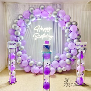 May include: A birthday party setup featuring a large balloon arch in shades of purple and silver. A neon sign reads "Happy Birthday". The scene includes clear pedestals with balloons and a cake, creating a festive atmosphere.