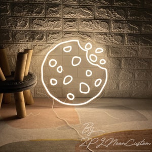 May include: White neon sign shaped like a cookie with a bite taken out of it. The sign is lit up and has a warm glow. The sign is on a light brown rug with a white cord.