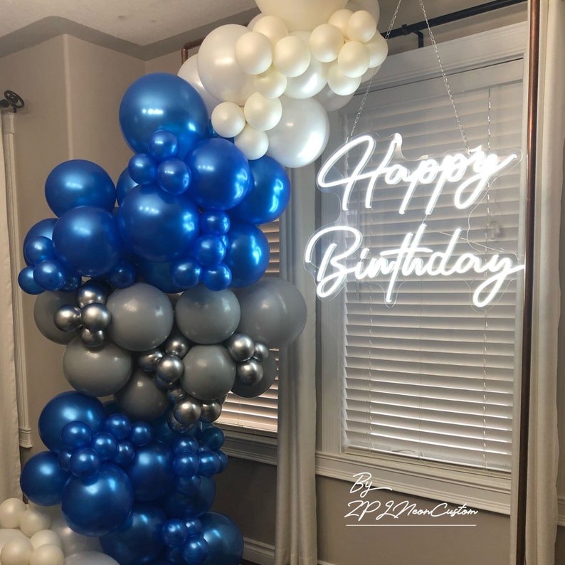May include: A balloon garland in shades of blue, silver, and white, with a neon sign that reads "Happy Birthday." The balloons are arranged in a cascading design, perfect for a birthday celebration.