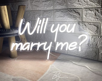 May include: White neon sign with the text "Will you marry me?" on a clear acrylic background. The sign is sitting on a light brown rug in front of a brick wall.