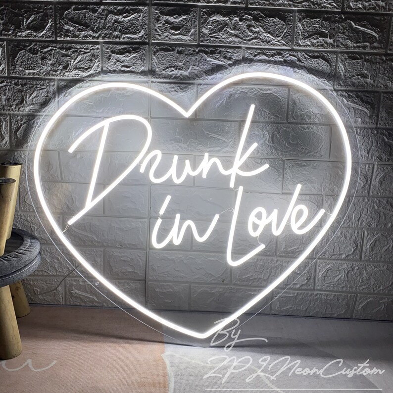 May include: A white neon sign in the shape of a heart, with the words "Drunk in Love" written in cursive script. The sign is against a white brick wall. The sign is made by ZPZL Neon Custom.