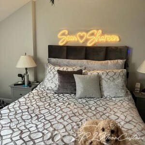 May include: A warm yellow neon sign mounted above a headboard, displaying the names Sean and Shareen separated by a heart symbol in a cursive script font. The sign emits a soft glow against the wall, illuminating the bedroom space below.