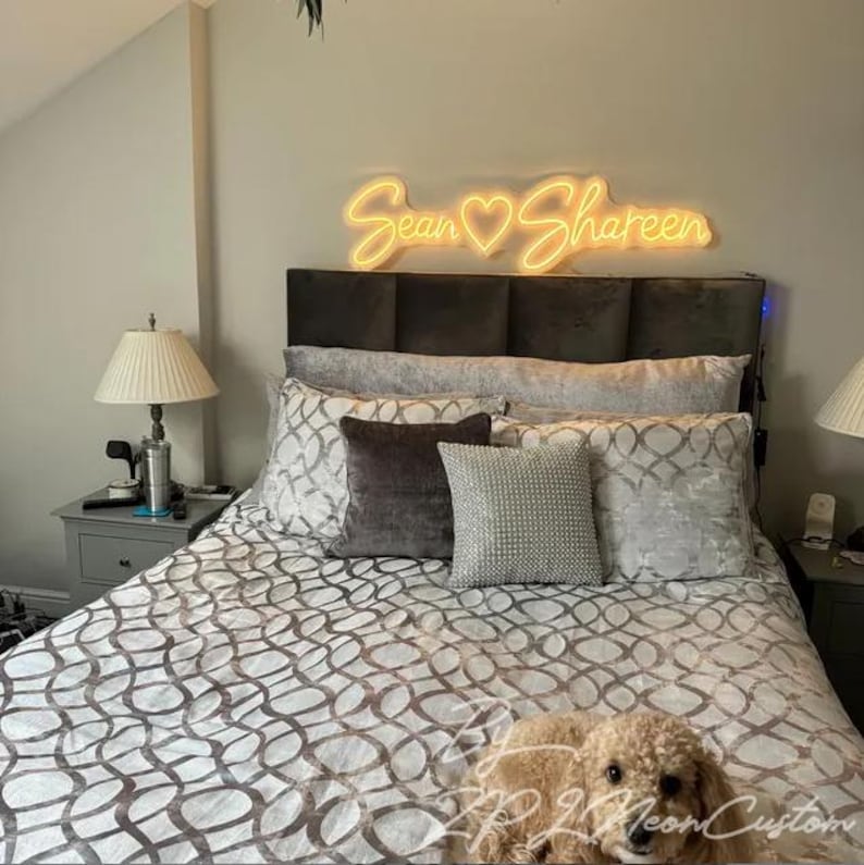 May include: A warm yellow neon sign mounted above a headboard, displaying the names Sean and Shareen separated by a heart symbol in a cursive script font. The sign emits a soft glow against the wall, illuminating the bedroom space below.