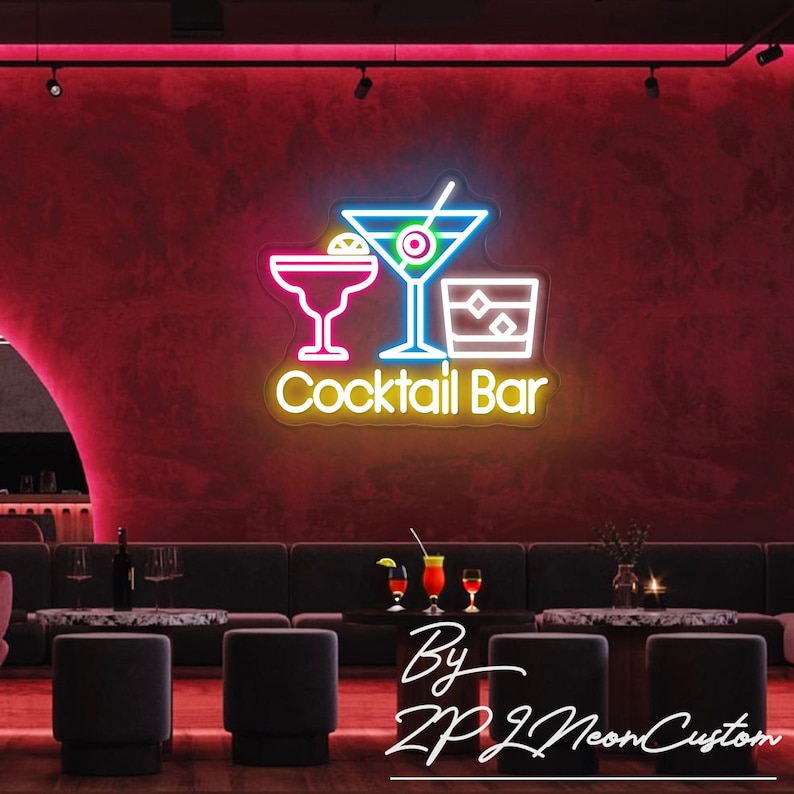 May include: A neon sign for a cocktail bar, featuring three cocktail glasses in white, pink, and blue, with the words "Cocktail Bar" in yellow. The sign is mounted on a red wall, with a bar setting in the background.