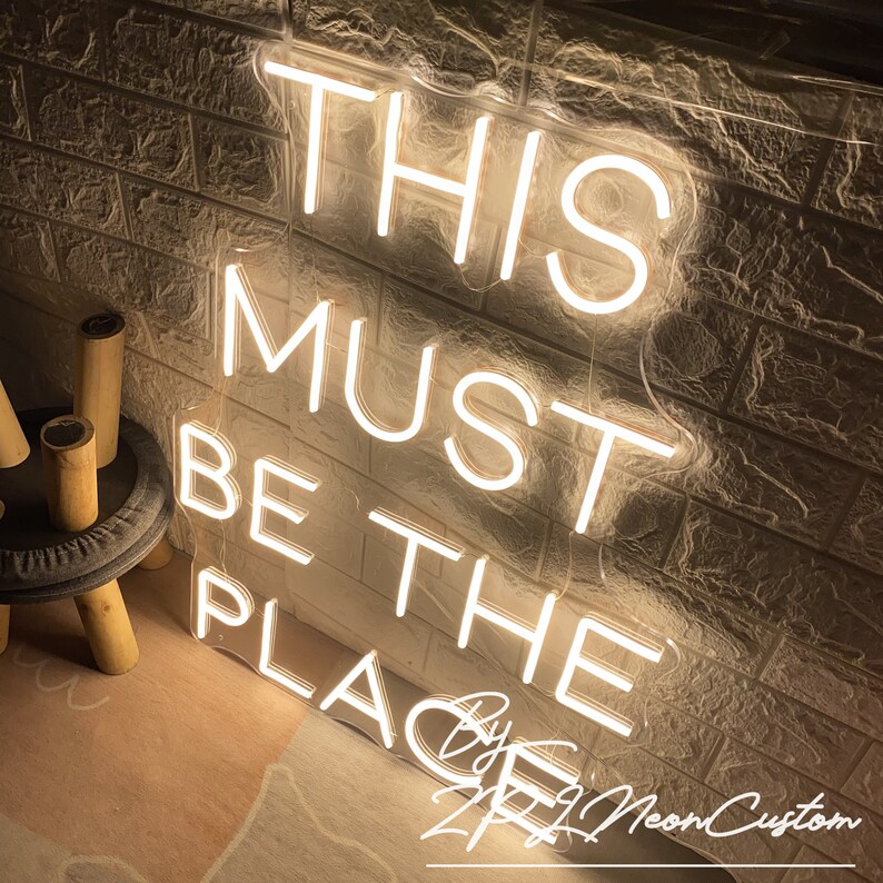 Op de afbeelding: Een neonbord met de tekst "THIS MUST BE THE PLACE" in een heldere, warme witte kleur. Het bord is bevestigd aan een getextureerde, lichtgekleurde bakstenen muur. De tekst "By ZZZ NeonCustom" is ook aanwezig.