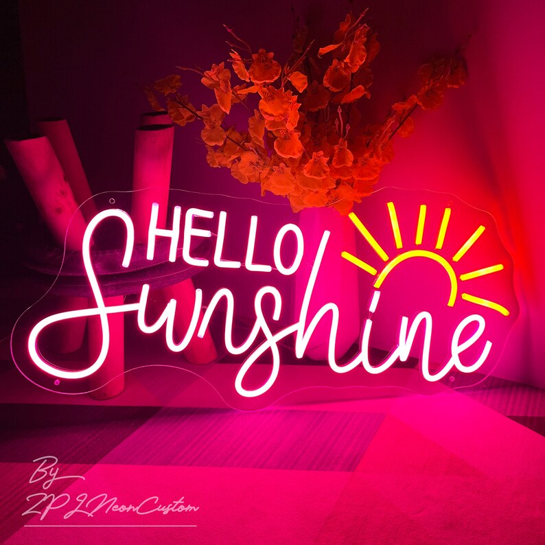 May include: A neon sign in a cursive font that reads "HELLO Sunshine" with a yellow sun graphic. The sign is outlined in white and glows with a pink light. The background is a dark pink.