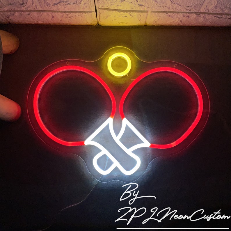 May include: A neon sign in the shape of two ping pong paddles with a white and red glow. A yellow circle is centered above the paddles. The text "By ZPL Neon Custom" is written in white cursive.
