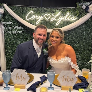 May include: A custom warm white neon sign featuring the names Cory and Lydia in a script font, measuring 39.5 inches wide. The sign is mounted on a green hedge wall backdrop decorated with white and blue floral arrangements and draped fabric.
