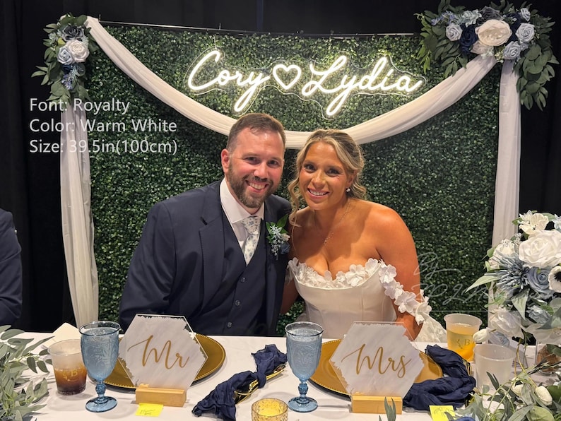 May include: A custom warm white neon sign featuring the names Cory and Lydia in a script font, measuring 39.5 inches wide. The sign is mounted on a green hedge wall backdrop decorated with white and blue floral arrangements and draped fabric.