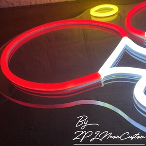 May include: Close-up of a neon sign with a red, white, and yellow color scheme. The sign is in the shape of a flower. The text "By ZP Neon Custom" is written in white cursive font.