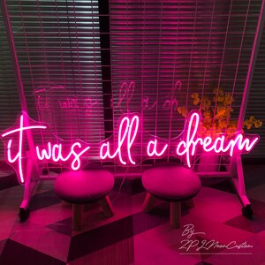 May include: A neon sign in bright pink script reads "It was all a dream." The sign is mounted on a white metal frame, with two small stools in front. The background includes a window with blinds and a small vase of yellow flowers.