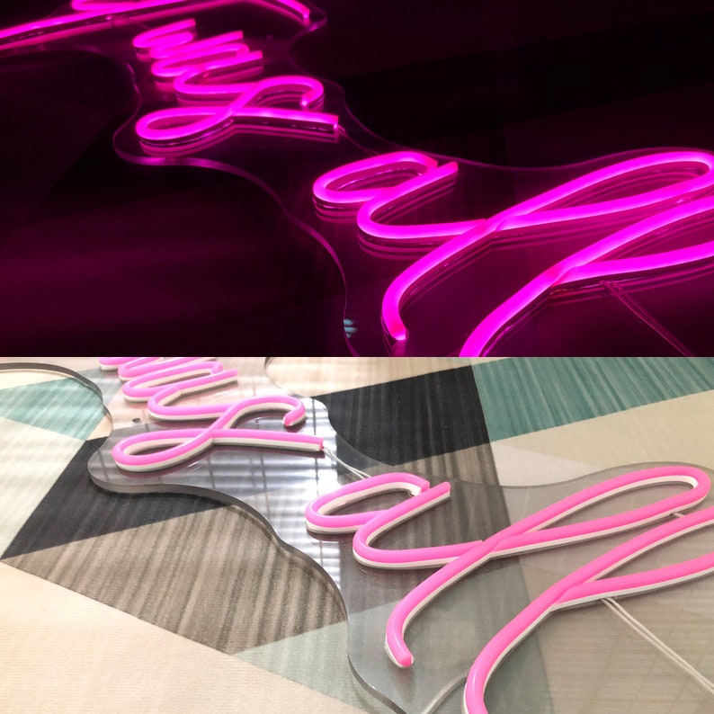 May include: A neon sign in a cursive font, illuminated in bright pink. The sign is mounted on a clear, irregular-shaped backing. The background transitions from black to a patterned surface.