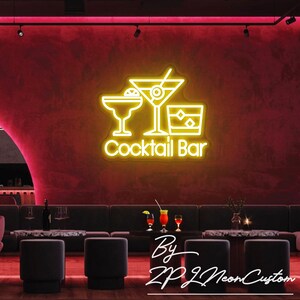 May include: A neon sign with the words "Cocktail Bar" and illustrations of a margarita, martini, and rocks glass. The sign is yellow and mounted on a red wall. The sign is in a bar setting with seating and tables.