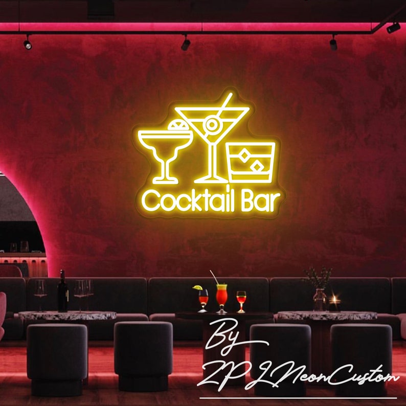 May include: A neon sign with the words "Cocktail Bar" and illustrations of a margarita, martini, and rocks glass. The sign is yellow and mounted on a red wall. The sign is in a bar setting with seating and tables.