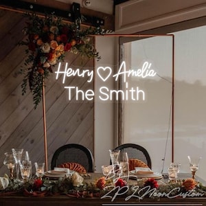 May include: A custom white neon sign featuring the names Henry and Amelia with a heart symbol in between, and the text The Smith below. The sign is mounted on a copper-colored metal frame decorated with a floral arrangement of red and orange flowers.