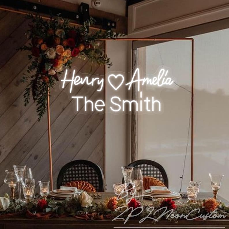 May include: A custom white neon sign featuring the names Henry and Amelia with a heart symbol in between, and the text The Smith below. The sign is mounted on a copper-colored metal frame decorated with a floral arrangement of red and orange flowers.