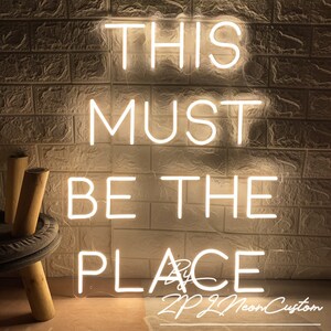 Op de afbeelding: Een wit neonbord met de tekst 'THIS MUST BE THE PLACE'. Het bord is bevestigd aan een getextureerde, gebroken witte bakstenen muur. De rechterbenedenhoek bevat de tekst 'By ZP ZL Neon Custom'.