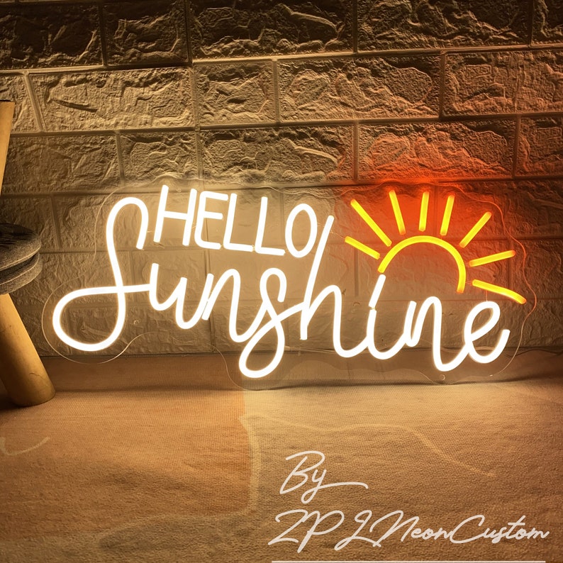 May include: A neon sign with the words "HELLO Sunshine" in white cursive script, with a yellow sun graphic. The sign is illuminated against a textured brick wall. The sign is made by ZPJ Neon Custom.