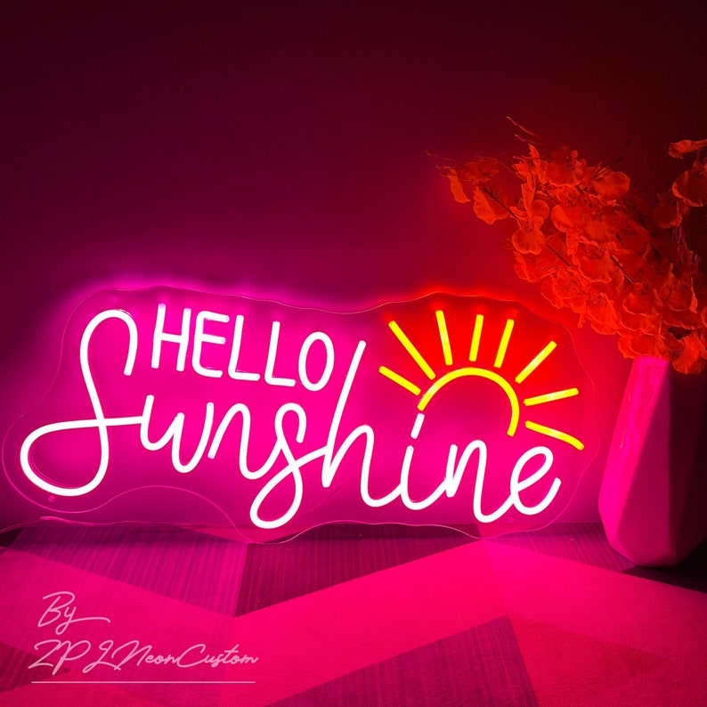 May include: A neon sign with the words "HELLO Sunshine" in white script, with a yellow sun graphic. The sign is encased in a pink outline and glows with a bright pink light. The sign is on a pink surface.