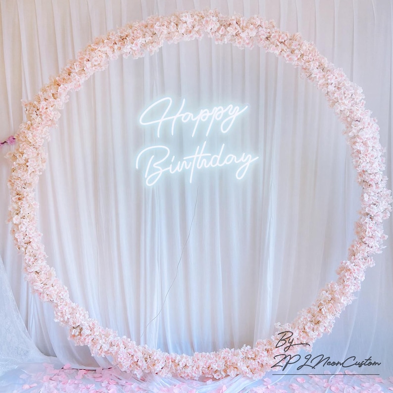 May include: A circular floral arrangement with light pink blossoms, surrounding a neon sign that reads "Happy Birthday." The backdrop is a white curtain, with scattered petals on the floor. The sign is a custom design.