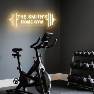 May include: A home gym setup featuring a neon sign with the text "THE SMITH'S HOME GYM" in a dumbbell shape. A black exercise bike with a red seat is in the foreground. Dumbbells are on a rack in the background. The walls are dark gray.