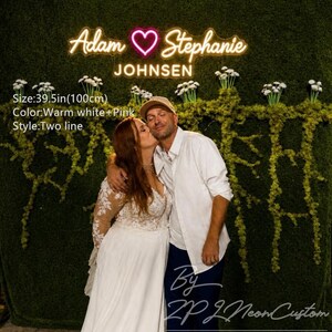 May include: A warm white neon sign featuring the text Adam, a heart symbol, and Stephanie, with the word JOHNSEN below. The sign is 100cm wide and displays in two lines against a green moss wall backdrop with white floral accents.