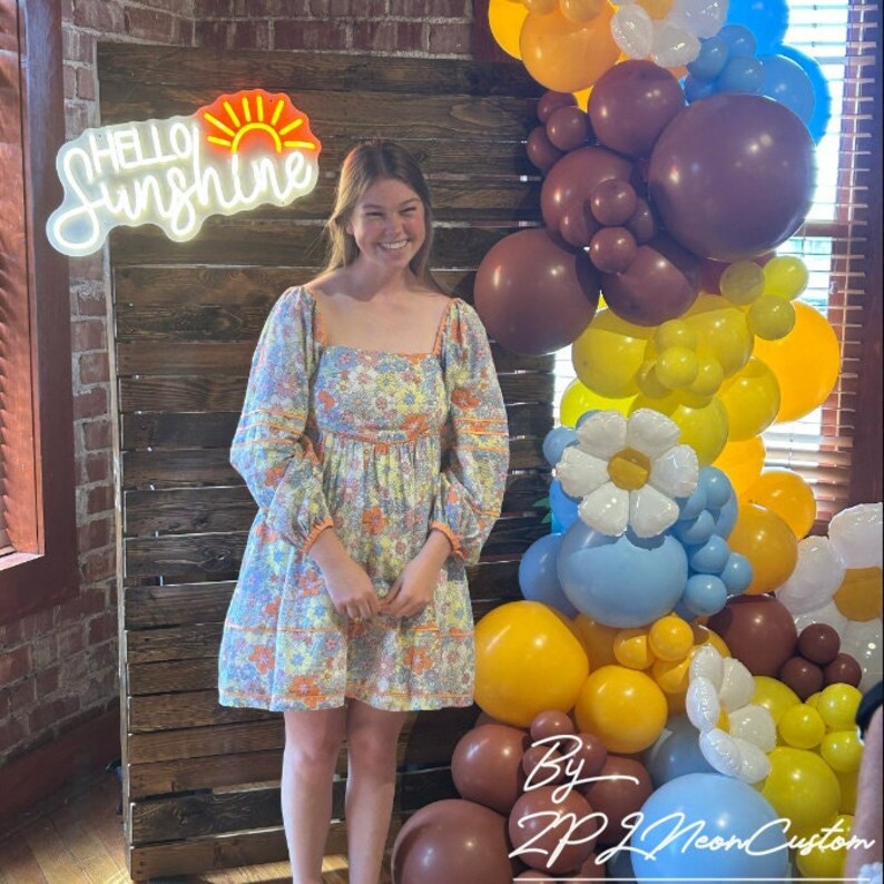 May include: A woman stands in front of a wooden backdrop, smiling. She wears a floral print dress with long sleeves. Behind her is a balloon arch in shades of yellow, brown, and blue, and a neon sign that reads "Hello Sunshine."