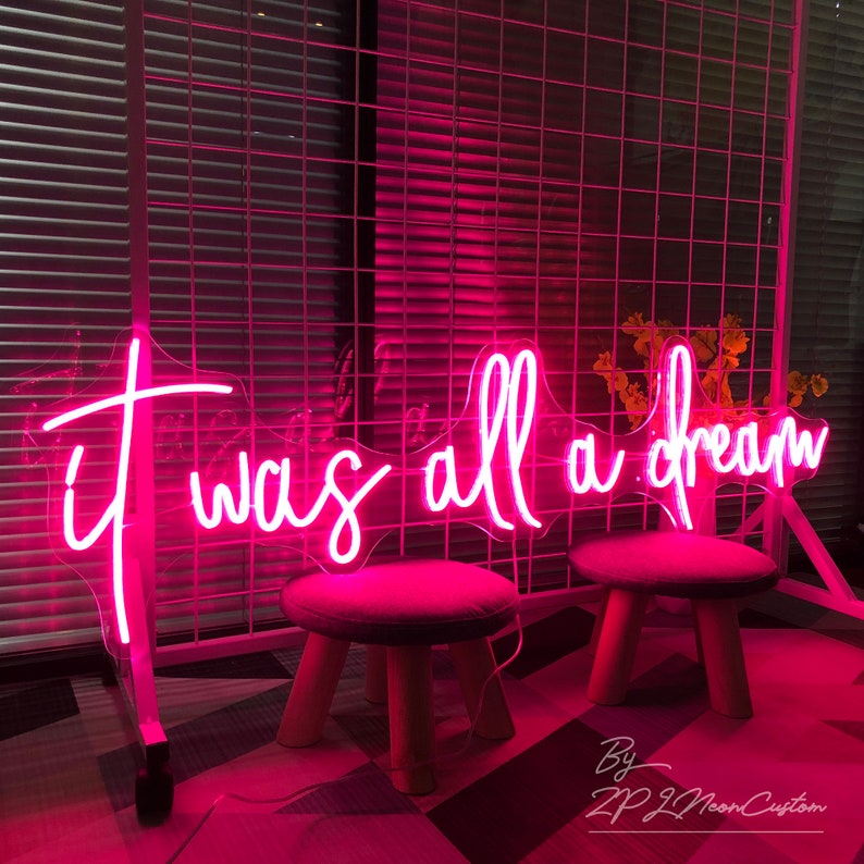 May include: A bright pink neon sign with the words "it was all a dream" in a cursive font. The sign is mounted on a white metal grid. Two small stools with wooden legs and pink seats are in front of the sign.