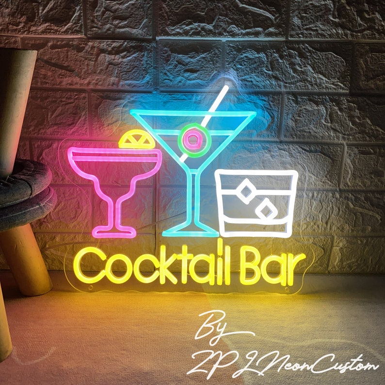 May include: Neon sign with the words "Cocktail Bar" in yellow, and three cocktail glass designs in pink, blue, and white. The sign is against a brick wall.