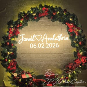 May include: A circular Christmas wreath made of green pine branches decorated with red and gold ornaments, red poinsettia flowers, and string lights. A white neon sign in the center displays the text Jannik ❤️ Annkathrin and 06.02.2026.