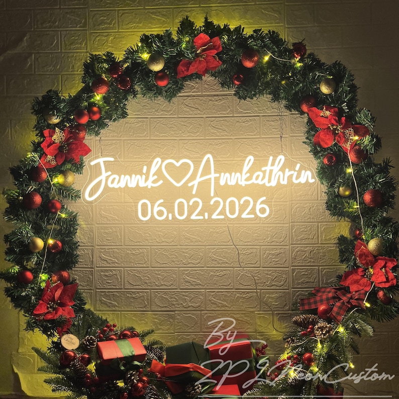 May include: A circular Christmas wreath made of green pine branches decorated with red and gold ornaments, red poinsettia flowers, and string lights. A white neon sign in the center displays the text Jannik ❤️ Annkathrin and 06.02.2026.