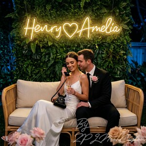May include: A glowing yellow neon sign featuring the names Henry and Amelia separated by a heart symbol. The script-style lettering is mounted against a lush green foliage wall, emitting a warm light that illuminates the surrounding area.