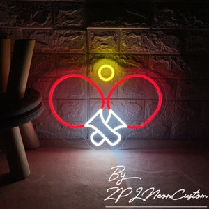May include: A neon sign featuring a ping pong paddle design with red outlines and white handles. A yellow ball is above the paddles. The sign is against a textured brick wall. The text "By ZP Neon Custom" is at the bottom.