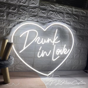 May include: A white neon sign in the shape of a heart with the words "Drunk in Love" in a cursive font. The sign is against a white brick wall. The sign also includes the text "By ZPJ Neon Custom".