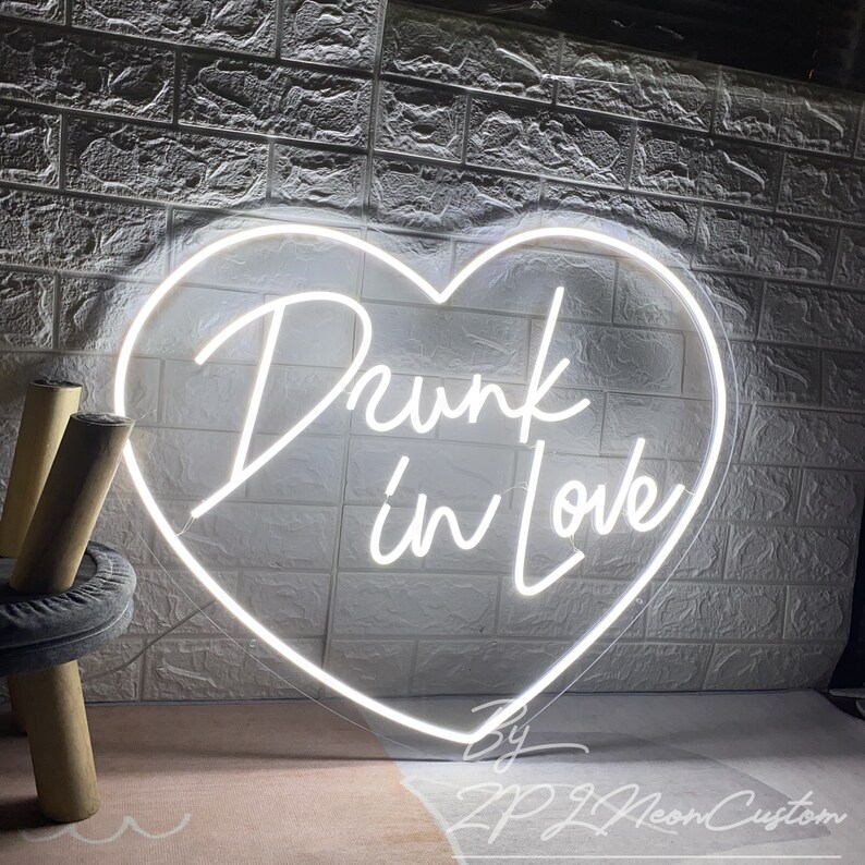 May include: A white neon sign in the shape of a heart with the words "Drunk in Love" in a cursive font. The sign is against a white brick wall. The sign also includes the text "By ZPJ Neon Custom".