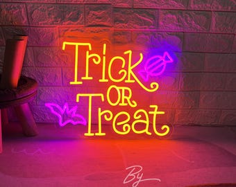 May include: A neon sign with the words "Trick or Treat" in yellow, with a purple bat and candy graphic. The sign is lit against a brick wall, with a gradient of pink and orange light.