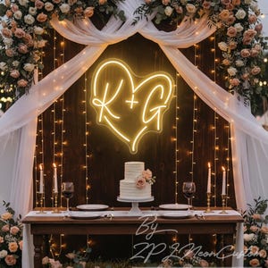May include: A wedding reception setup with a neon heart sign displaying the initials "R+G". A two-tiered cake with floral accents sits on a table set for a meal. The background features string lights, flowers, and draped fabric.