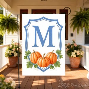 Peut inclure: Un drapeau de jardin rectangulaire présentant une grande lettre M bleue centrée dans un contour de bouclier bleu. Sous la lettre, trois citrouilles orange avec des feuilles vertes sont disposées sur un fond blanc uni.