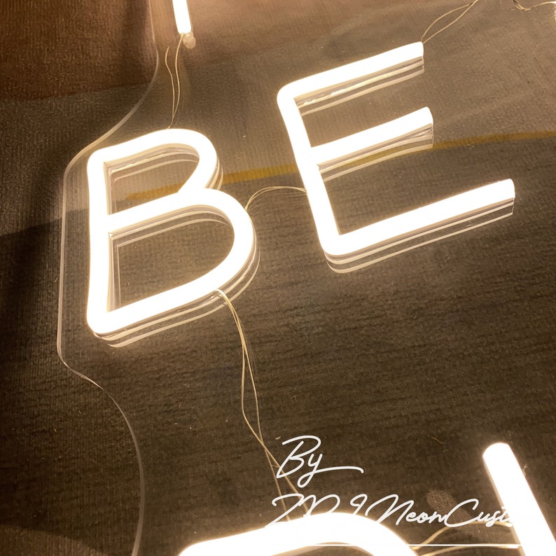 Op de afbeelding: Een close-up van een neonbord met het woord "BE". De letters zijn wit en verlicht, met een doorzichtige achterkant. Het bord staat op een donkere ondergrond. De tekst "By M9 NeonCusi" is ook zichtbaar.