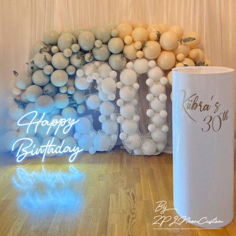 May include: A birthday party decoration featuring a neon sign that reads "Happy Birthday" in blue. The number "30" is formed with white balloons, and a white cylindrical stand displays "K&uuml;bra's 30th" in gold lettering. The background has a gradient of balloons.