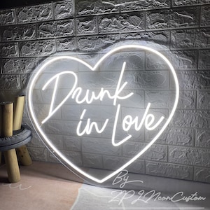 May include: A white neon sign in the shape of a heart, with the words "Drunk in Love" written in cursive. The sign is mounted on a textured, light gray brick wall. The sign is made by ZP22 Neon Custom.