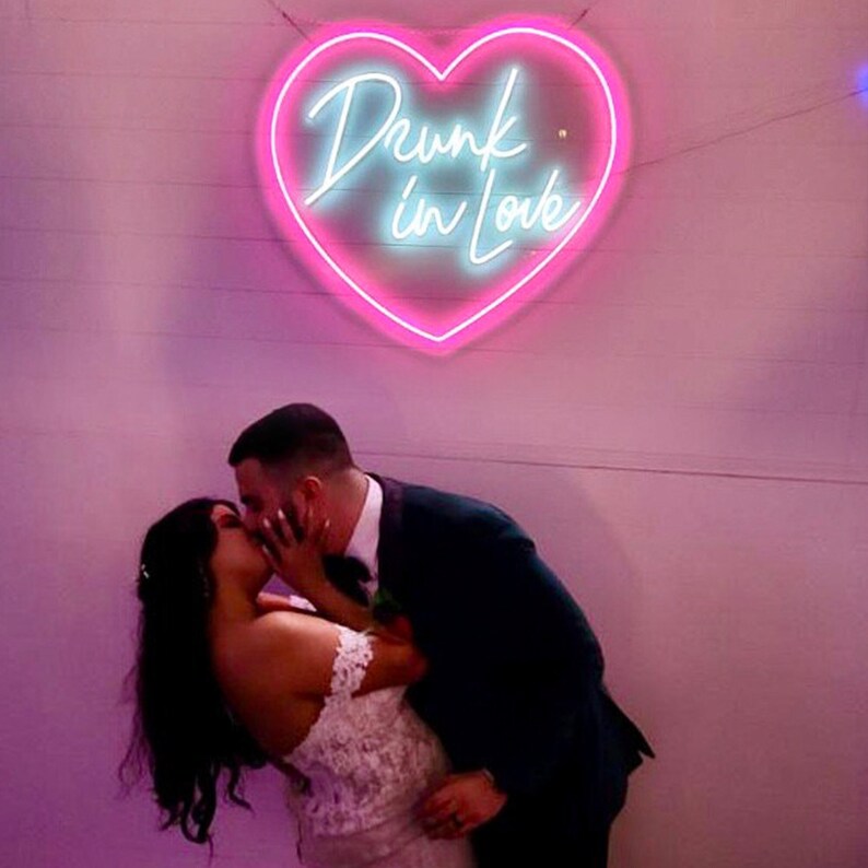 May include: A neon sign in the shape of a heart with the words "Drunk in Love" in cursive script. The sign is pink with a light blue inner glow. A couple is kissing in front of the sign.
